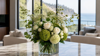 Luxury green and white faux floral arrangement creamy white roses, green hydrangeas, Queen Anne’s lace, airy botanical stems, and lush greenery styled in a modern clear glass vase on a marble table in an elegant coastal-inspired living room