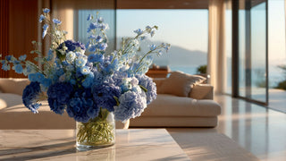 Blue hydrangea faux floral arrangement in a clear glass vase on a marble table, styled in a bright coastal modern living room with ocean view and soft sunlight