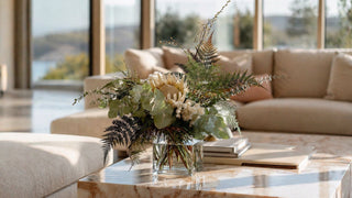 Luxury faux eucalyptus and mixed greenery arrangement in a clear glass vase on a marble coffee table, styled in a bright modern coastal living room with ocean view and soft natural sunlight, elegant high-end home decor