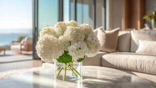 Luxury faux white hydrangea bouquet in a clear glass vase on a marble coffee table, styled in a bright coastal living room with ocean view and soft natural sunlight, elegant high-end home decor