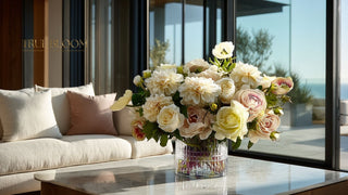 Soft neutral faux floral arrangement with cream and blush roses in a textured glass vase, styled in a modern living room with ocean view by True Bloom Floral