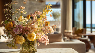 Yellow and blush wildflower faux floral arrangement with airy textures in a glass vase, styled on a marble coffee table in a bright modern living room with warm sunlight