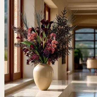 Black Norfolk pine artificial stems add dark architectural height to burgundy calla lilies, dusty-rose orchids and plum florals in an ivory vase, styled as a dramatic fall and winter arrangement in a sunlit modern home.