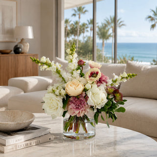 Blush Garden Manor True Bloom premade floral arrangement styled in a clear glass vase on a marble coffee table, featuring white and cream roses, blush dahlias, white snapdragon-style stems, soft mauve anemone-style blooms, greenery, and real touch florals in a bright coastal living room.