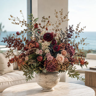 Burgundy Faux Dried Pomegranate Branch styled in a large fall floral arrangement with burgundy dahlias, mauve roses, dried hydrangeas, ivory filler stems, autumn foliage, and layered greenery in a stone vessel on a marble coffee table in a bright coastal living room.