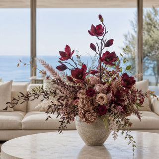 Burgundy Magnolia Tree Stem styled as a sculptural focal stem in a large fall floral arrangement with mauve roses, burgundy blooms, dried hydrangeas, airy filler stems, and seasonal greenery in a stone vessel on a marble coffee table in a bright coastal living room.