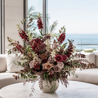 Burgundy Stock Stem styled as a tall movement flower in a fall floral arrangement with mauve roses, burgundy blooms, airy filler stems, dried-style branches, and layered greenery in a stone vessel on a marble coffee table in a bright coastal living room.