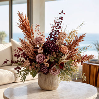 Burnt Terracotta Sorghum Grass Stem styled in a fall floral arrangement with mauve roses, burgundy blooms, dried-style filler stems, eucalyptus greenery, and warm terracotta foliage in a textured ceramic vase on a marble coffee table in a bright coastal living room.