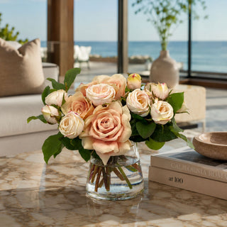 Champagne Rose Manor True Bloom Real Touch premade floral arrangement styled in a clear glass vase on a marble coffee table with champagne roses, cream rose buds, green foliage, and soft neutral florals in a bright coastal living room.