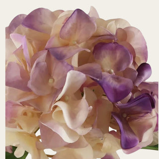 Close-up of the 20 inch real touch purple and cream faux hydrangea stem showing the green stem, textured faux leaf, and lower hydrangea petals in soft cream, blush-lilac, and purple tones against a warm cream background.