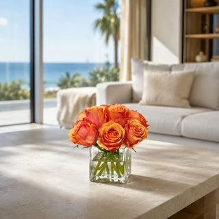 Coral Rose faux floral premade arrangement styled in a clear glass square vase on a stone coffee table with warm coral, peach, and orange rose blooms in a bright coastal living room.
