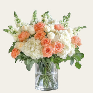 Coral True Bloom duchess roses styled with white hydrangeas, cream roses, and greenery in a clear glass vase for a warm refined faux floral arrangement.