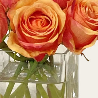 Close up view of the Coral Rose faux floral premade arrangement shown in a clear glass square vase with compact coral, peach, and orange rose blooms for coffee table, bedside table, bathroom vanity, kitchen shelf, and small console styling.