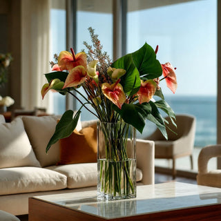 Coral and Green Anthurium stems styled with lush green calla leaves and textural greenery in a clear glass vase on a marble coffee table in a luxury coastal living room; ideal for spring, summer, and winter arrangements because the sculptural anthurium blooms create focal interest and movement while the bold foliage adds depth and balance