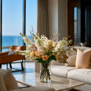 Cream lilac stems styled in a large clear glass vase arrangement on a marble coffee table in a luxury coastal living room with ocean views, cream seating, peach accents, and soft natural light.