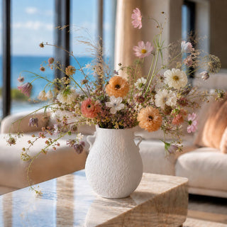 Airy early fall floral arrangement in a matte white textured vase with side handles, styled on a marble coffee table in a soft coastal living room, featuring peach, blush, ivory, and muted mauve blooms with delicate wispy stems and natural sunlight.