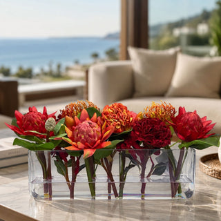 Ember Reserve faux floral premade arrangement styled in a clear glass vase on a marble coffee table with red protea-style blooms, ember orange florals, coral accents, green foliage, and warm seasonal color in a bright coastal living room.