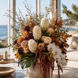 Decorative floral arrangement featuring fall inspired florals and the white king flocked protea stem with autumnal colors in a vase, set against a scenic background.