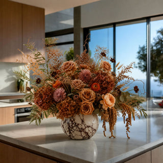 A large dried floral arrangement in a white marble-patterned vase, placed on a light stone kitchen island with a modern kitchen and large windows in the background.