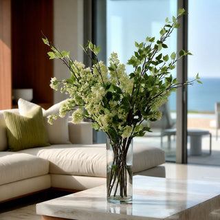 Faux46 inch green leaf stem styled in a clear glass vase with white budding flower stems. place don a marble coffee table of a luxury coastal view living room 