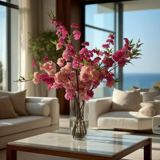 Fuchsia faux cherry blossom spray stems styled with soft pink cherry blossoms in a clear glass vase on a marble coffee table in a luxury coastal living room; ideal for spring and summer arrangements because the vibrant fuchsia blooms create focal interest and movement while the softer pink blossoms add depth, balance, and a layered garden-inspired look.