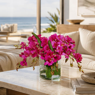 Fuchsia Conservatory faux floral premade arrangement styled in a clear glass vase on a marble coffee table with vivid fuchsia orchid-style blooms, green foliage, and a wide low silhouette in a bright coastal living room.
