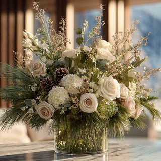 Neutral winter floral arrangement with cream roses, hydrangeas, pine greenery and pinecones in a clear glass vase styled on a marble table in a cozy sunlit interior