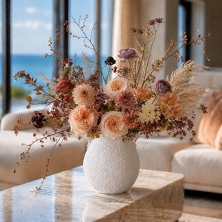 Airy late fall floral arrangement in a matte white textured vase with side handles, styled on a marble coffee table in a soft coastal living room, featuring peach, blush, ivory, and muted mauve blooms with delicate wispy stems and natural sunlight.