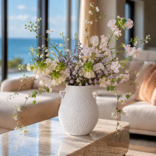 Matte white textured vase styled on a marble table in a coastal modern living room with an airy faux floral arrangement of soft lilac lily of the valley, blush-white sweet peas, pale cream fritillaria, and delicate flowering jasmine vine for refined spring and summer home decor.