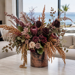 Mustard Amaranthus Stem styled in a copper vase arrangement with burgundy roses, mauve florals, protea-style blooms, eucalyptus, and layered fall greenery on a marble coffee table in a bright coastal living room.