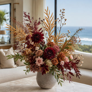 Orange and Tan Seeded EVA Leaves Stem styled as tall movement foliage in a fall floral arrangement with burgundy dahlias, rust roses, blush accents, cream filler flowers, burgundy leaves, and layered seasonal greenery in a stone vase on a marble coffee table in a bright coastal living room.