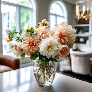 Peach Real Touch dahlia stems styled in a glass vase with cream dahlias, soft peach roses, ivory blooms, greenery, and airy branch texture in a bright living room arrangement for spring, summer, fall, and winter styling.
