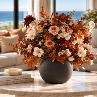 Small Black Konos Vase styled with a full fall faux floral arrangement featuring cream blooms, rust foliage, peach roses, burgundy dahlias, orange seasonal stems, and warm autumn greenery on a marble coffee table in a bright coastal-inspired living room.
