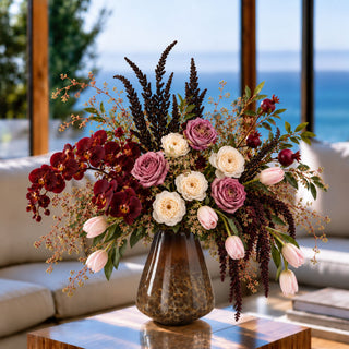 Small brown and cream blown glass vase holding burgundy orchids, mauve and cream flowers, pink tulips, and trailing amaranthus