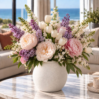 Small White Konos Vase styled with a full spring faux floral arrangement featuring soft pink peonies, white tulips, purple lilac-style stems, white filler flowers, and greenery on a marble coffee table in a bright coastal-inspired living room.