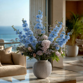 Blue True Bloom Real Touch delphinium stems styled in a white vase with layered neutral florals and airy greenery for a calm garden-inspired floral arrangement that works beautifully year-round because dusty blue tones pair naturally with both seasonal greenery and soft transitional palettes.
