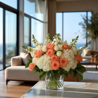 Luxury floral arrangement featuring white hydrangeas, peach roses, ivory spray roses, and white snapdragons styled in a clear glass vase on a marble coffee table in a modern coastal living room. Soft natural sunlight streams through floor-to-ceiling windows, creating an elegant designer-inspired interior with fresh floral centerpiece styling.