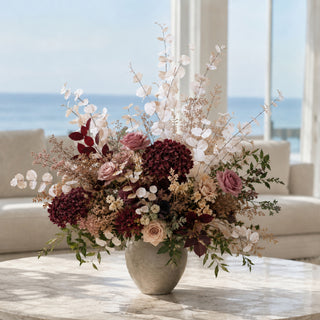 A large, elegant dried and fresh floral arrangement in a textured gray ceramic vase, placed on a marble table in a bright living room with a view of the ocean in the background.