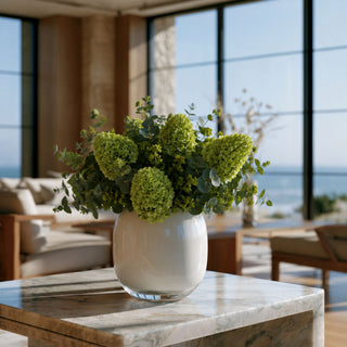 Luxury faux floral arrangement featuring green cone hydrangeas, eucalyptus, and soft green botanical accents styled in a glossy white glass vase on a marble coffee table in a modern coastal living room with ocean views. 