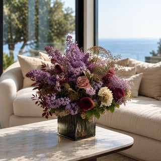 A large, vibrant purple and burgundy floral arrangement in a ribbed square glass vase, displayed on a white marble coffee table in front of a beige sofa with a window view of the ocean.