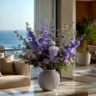 Purple True Bloom Real Touch delphinium stems styled with layered lavender florals and airy greenery in a textured vase for a romantic garden-inspired arrangement that works beautifully year-round because soft purple tones blend naturally with both seasonal greenery and transitional floral palettes.