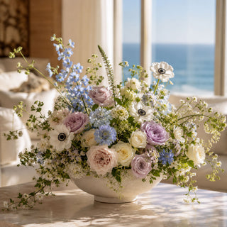 White real touch buttercup stems styled in a tall glass vase with pine greenery and creamy florals on a coffee table in a quiet luxury coastal living room