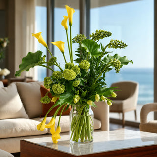 A vibrant floral arrangement featuring yellow calla lilies and green foliage in a clear glass vase, set on a marble-topped table in a modern living room with a window view of the ocean.