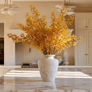 A large cream and brown stoneware vase filled with golden-yellow artificial yellow cimicifuga stems, placed on a marble kitchen island with a blurred modern kitchen in the background.