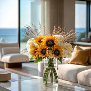 A vibrant artificial yellow sunflower and white flower bouquet in a clear glass vase, styled on a marble coffee table in a bright, modern living room with a blurred ocean view background.