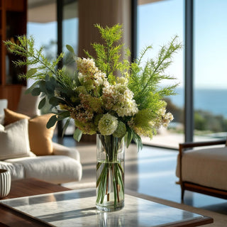Large floral arrangement styled with asparagus fern bush stems and other greenery on a marble coffee tale in a modern living room with ocean views in the background.