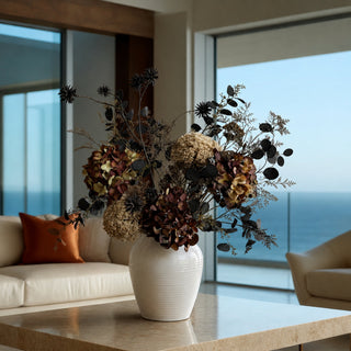 A dried floral arrangement in a ribbed white ceramic vase, placed on a light stone table in a modern living room with a large window overlooking the ocean.