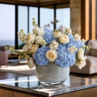 A floral arrangement in a round white vase, featuring light blue hydrangeas and cream roses, displayed on a polished marble table in a modern, sunlit living room.