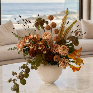 Large autumn floral arrangement in warm rust, caramel, cream and olive tones styled in an ivory textured vase on a marble table in a luxury coastal living room with ocean views.