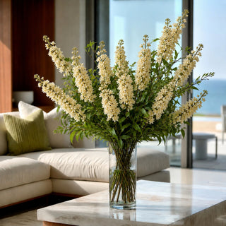 A large floral arrangement of cream-white blossoms with green foliage in a clear glass vase, placed on a marble coffee table in a bright modern living room with a sofa and large windows.